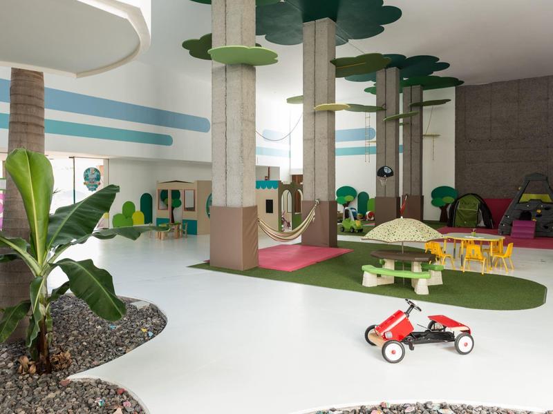 Aire de jeux intérieure lumineuse avec grandes décorations d'arbres, toboggans et jouets d'enfants sur un sol blanc.