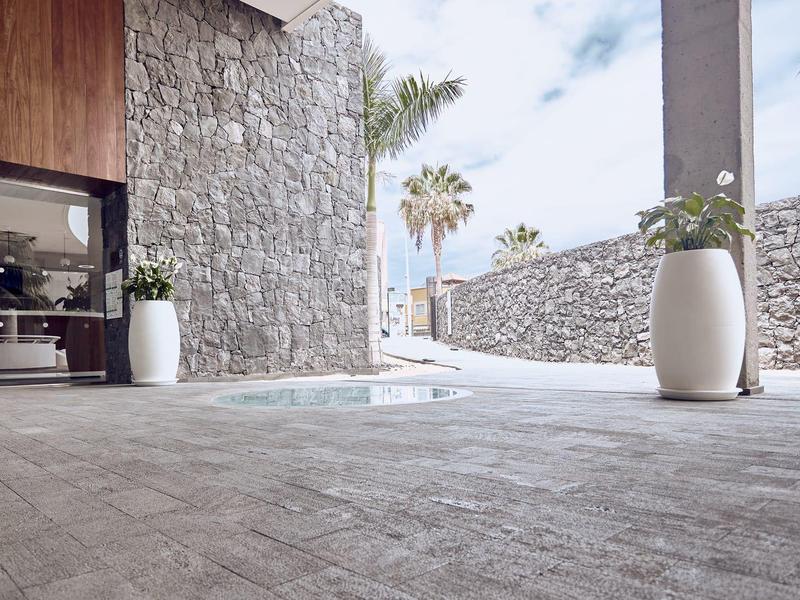 Espace piscine d'hôtel moderne avec murs en pierre, grands pots blancs et palmiers en arrière-plan.