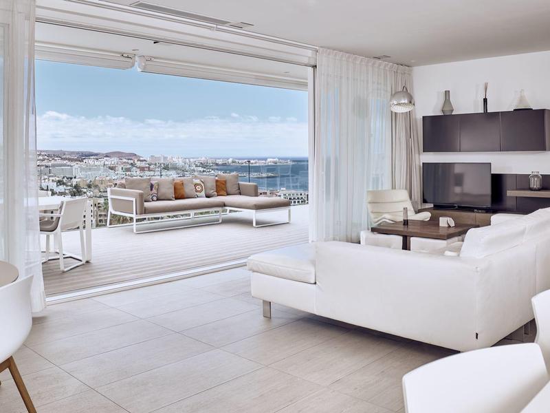 Espace de vie lumineux et moderne avec mobilier blanc et vue sur la mer et la ville depuis la terrasse.