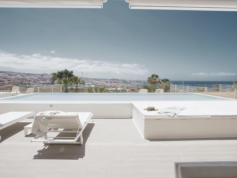 Espace piscine moderne d'hôtel avec chaises longues et vue sur la mer et la ville.