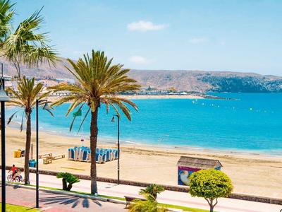 Strand mit blauem Meer, Palmen, Sand und Bergen im Hintergrund bei klarem Himmel.
