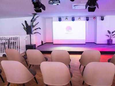 Nowoczesna sala konferencyjna z krzesłami, sceną i ekranem projekcyjnym oświetlona kolorowym światłem.