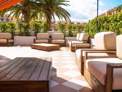 Offene Terrasse mit Holzmöbeln, weiß-beigem Bodenmuster und grünen Pflanzen im Hintergrund.