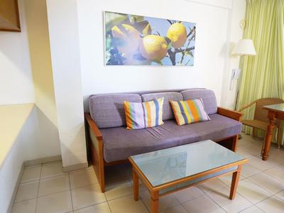 Kleines Wohnzimmer mit grauem Sofa, bunten Kissen, Glas-Couchtisch und großem Obstbild an der Wand.