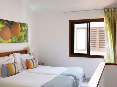 Helles Zimmer mit zwei Einzelbetten, bunten Kissen, Fruchtbild an der Wand und grünem Vorhang am Fenster.