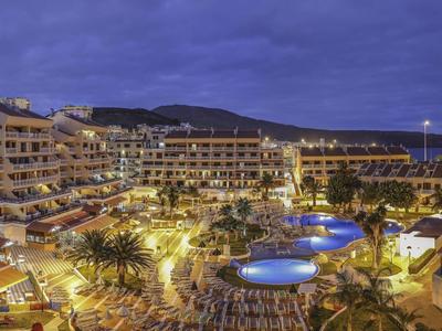 Hotel mit beleuchteten Pools, Palmen und Liegestühlen unter einem blauen Abendhimmel.