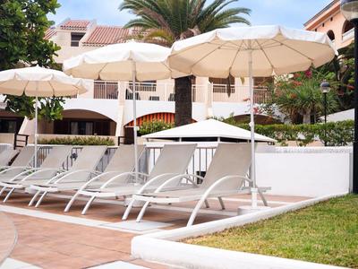 Weiße Liegestühle unter großen weißen Sonnenschirmen neben einem Hotel mit Palmen und grünem Rasen.