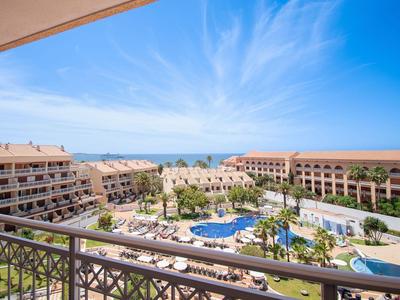 Widok z balkonu na kompleks hotelowy z basenami i palmami pod błękitnym niebem.