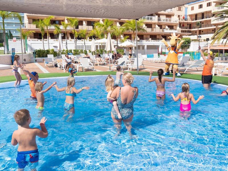 Kinder und Erwachsene spielen und planschen gemeinsam in einem flachen Hotelpool bei Sonnenschein.