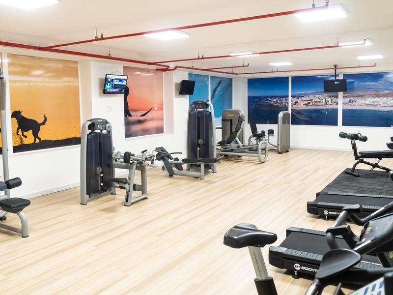 Heller Fitnessraum mit Cardio- und Kraftgeräten, großen Fenstern und hellem Holzboden.