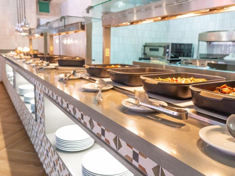 Langes Buffet mit warmen Speisen in Edelstahlbehältern und Tellern darunter in heller Umgebung.