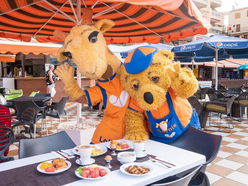 Zwei Personen in Tierkostümen sitzen an Außentisch mit Tellern voller Essen in sonnigem Café.