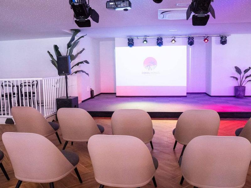 Ein heller Seminarraum mit rosa Stühlen, einer Bühne und zwei großen Pflanzen.