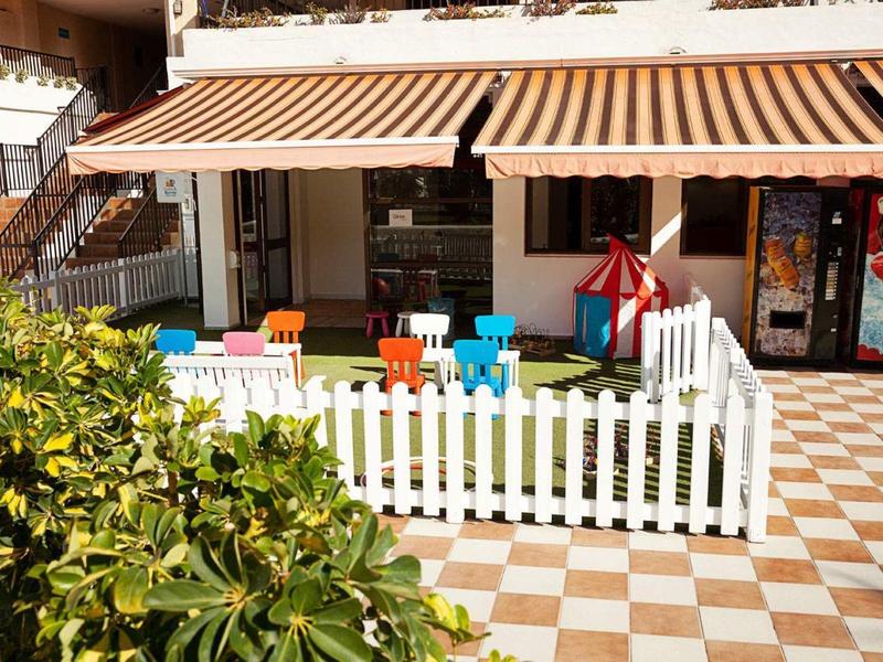 Kinderspielplatz mit bunten Stühlen und Tisch, weißem Zaun und Markise in warmem Licht.