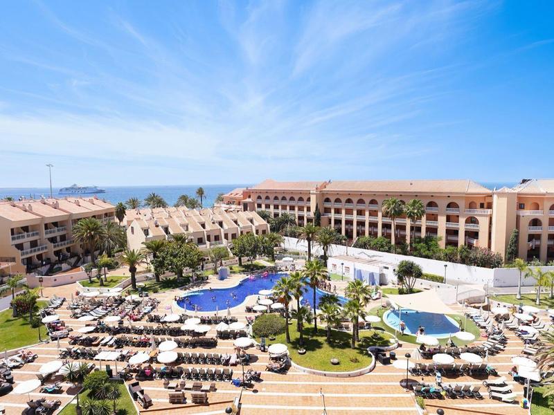 Große Hotelanlage mit Pools, Sonnenliegen und Palmen unter blauem Himmel mit leichter Bewölkung.
