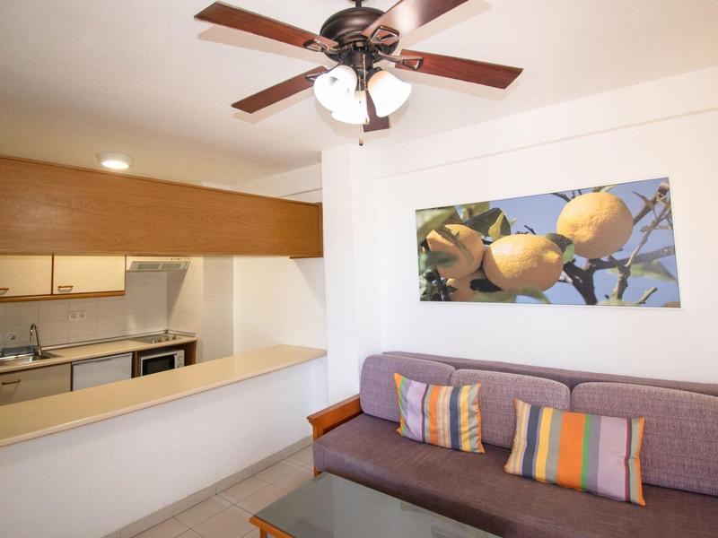 Wohnzimmer mit violettem Sofa, bunten Kissen, Deckenventilator und Küchenzeile im Hintergrund.