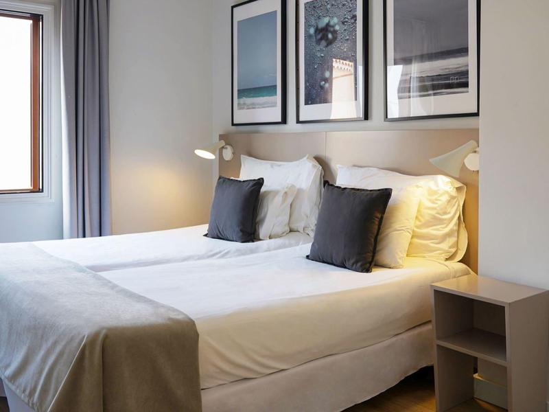 Ein helles Schlafzimmer mit zwei Einzelbetten, weißen Bettlaken, grauem Vorhang und Wandbildern.