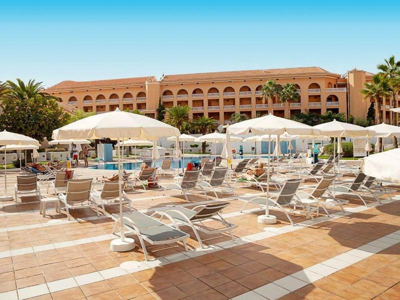 Großer Poolbereich mit weißen Liegestühlen und Sonnenschirmen vor einem Hotel mit roten Ziegeln.