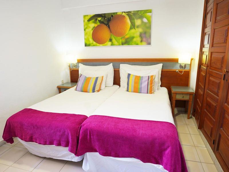 Gemütliches Doppelbett mit weißen Kissen, pinker Decke und Obst-Bild an weißer Wand.