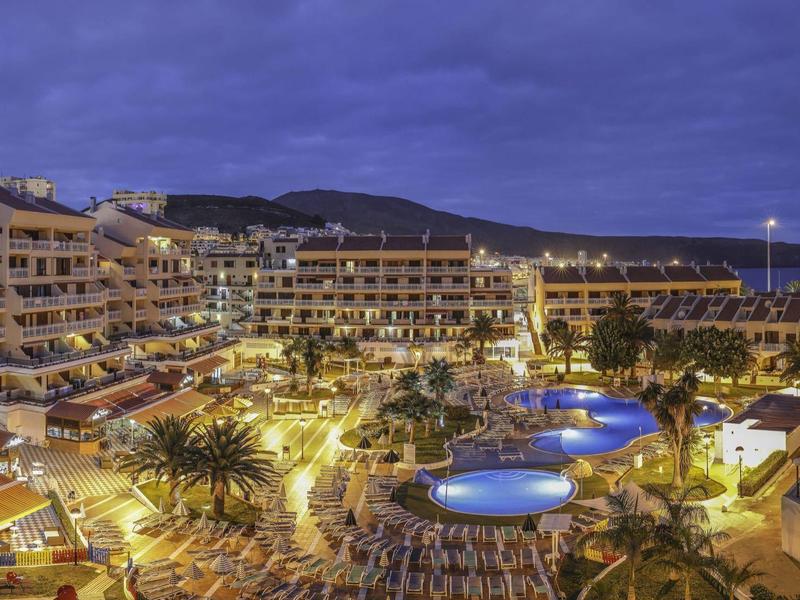 Hotel mit beleuchteten Pools, Palmen und Liegestühlen unter einem blauen Abendhimmel.