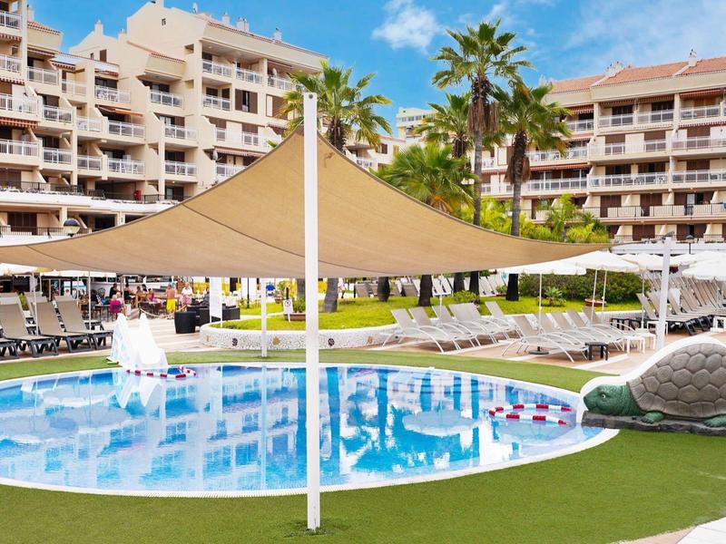 Kleiner Pool mit Sonnensegel, Liegestühlen und Hotelgebäuden im Hintergrund bei sonnigem Himmel.