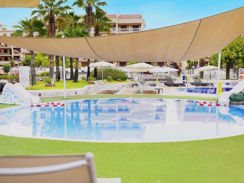 Runder Pool mit Sonnensegel, Liegestühlen und Palmen auf grüner Rasenfläche vor Hotelgebäuden.