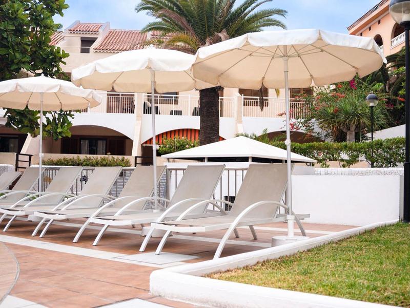 Weiße Liegestühle unter großen weißen Sonnenschirmen neben einem Hotel mit Palmen und grünem Rasen.