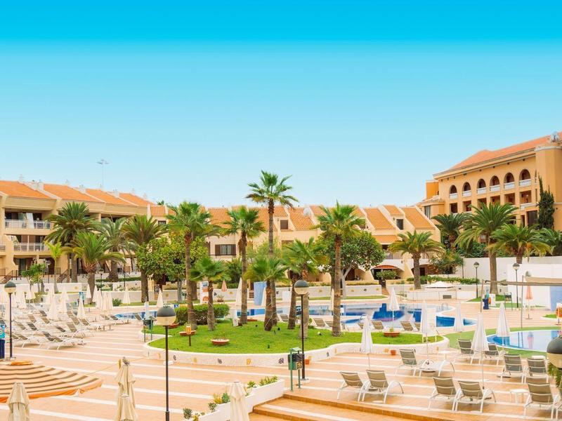 Großer Hotelkomplex mit mehreren Gebäuden, Pool, Palmen und Sonnenschirmen bei klarem Himmel.