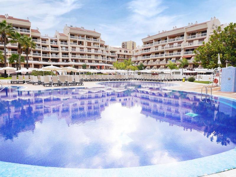 Großer blauer Pool mit mehreren Liegen und modernen Hotelgebäuden im Hintergrund bei Tageslicht.