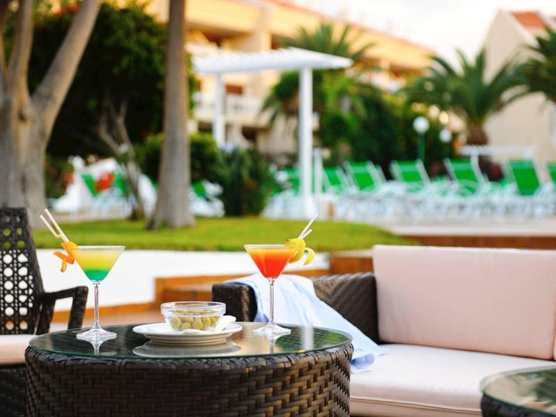 Outdoor-Lounge mit Tisch, zwei bunten Cocktails, Sofa und grüner Gartenlandschaft im Hintergrund.