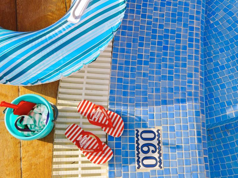 Blaue Fliesen am Pool, rote Sandalen, grüner Eimer mit Schaufel und blau-weiß gestreifter Schwimmreifen.