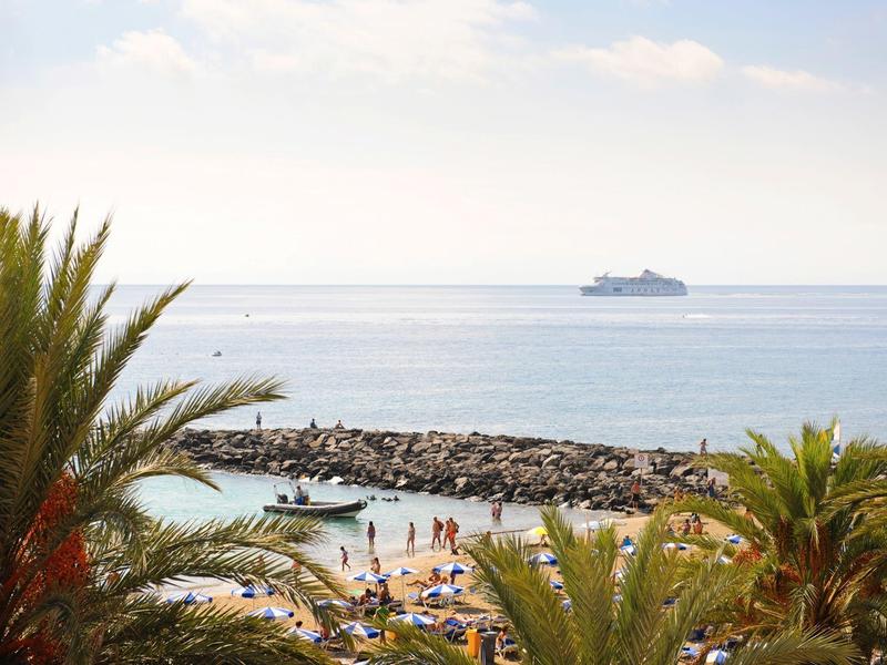 Strand mit Palmen, vielen Sonnenliegen, Menschen und einem Boot vor einer Felsenmole im Meer.