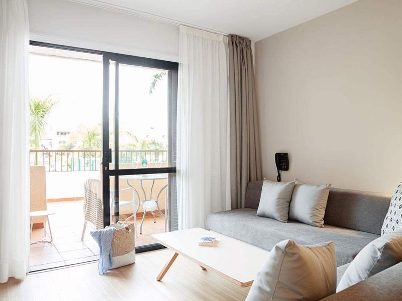 Helles Wohnzimmer mit grauem Sofa, Holztisch und großer Glas-Schiebetür zum Balkon mit Pflanzen.