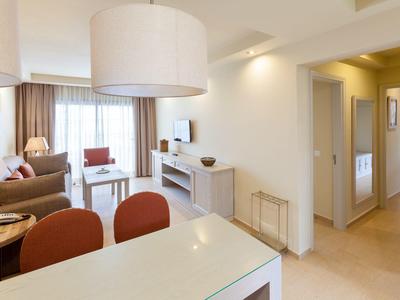 Helles Wohnzimmer mit weißen Wänden, beigem Boden, Sofa, Esstisch und großem Deckenlampenschirm.