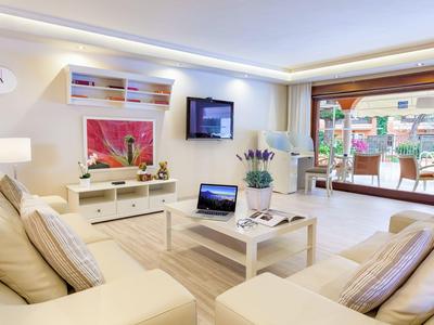 Heller Wohnraum mit beigen Sofas, weißem Tisch, TV, Kunstwerk und Zugang zur Terrasse mit Pflanzen.