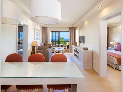 Helles Wohnzimmer mit Esstisch, roten Stühlen, Sofa und Blick auf Balkon mit Meer im Hintergrund.