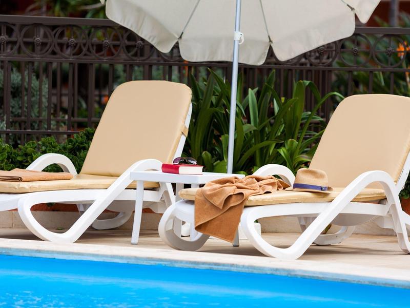 Zwei weiße Poolliegen mit Bezügen und Handtüchern neben blauem Wasser und grünem Pflanzenhintergrund.