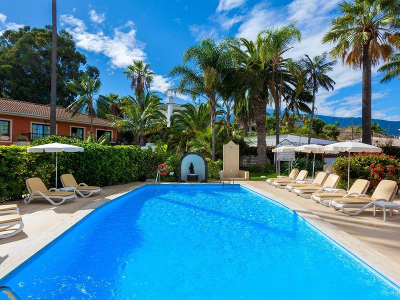 Schwimmbecken mit klarem blauem Wasser, umgeben von Liegestühlen und Palmen unter blauem Himmel.