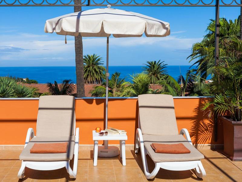 Zwei Liegestühle mit Sonnenschirm auf Terrasse, Meer und blauem Himmel im Hintergrund.