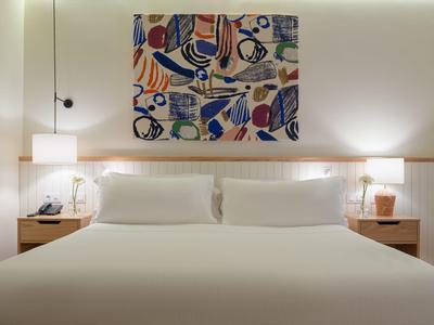 Nowoczesny pokój hotelowy z dużym łóżkiem podwójnym, dwoma stolikami nocnymi, lampami i kolorową abstrakcyjną sztuką na ścianie.