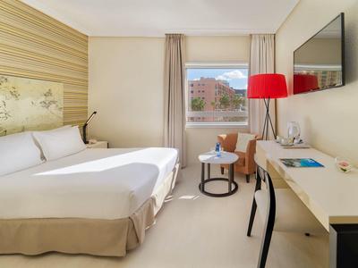 Jasny pokój hotelowy z łóżkiem podwójnym, biurkiem, czerwoną lampą i widokiem przez okno.