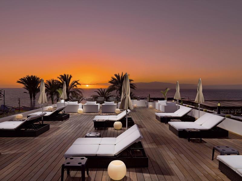 Taras na dachu hotelu z leżakami i palmami o zachodzie słońca nad morzem.