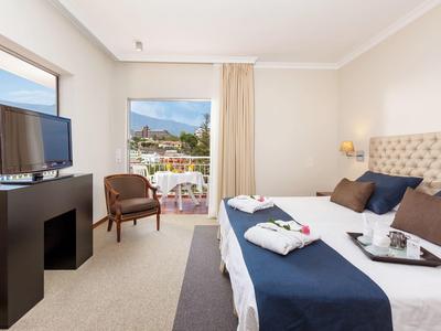 Helles Schlafzimmer mit großem Bett, Flachbildfernseher, Stuhl und Blick auf Balkon und Berge.