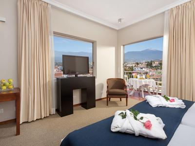 Helles Hotelzimmer mit zwei Betten, TV, Sessel und großen Fenstern mit Bergblick.