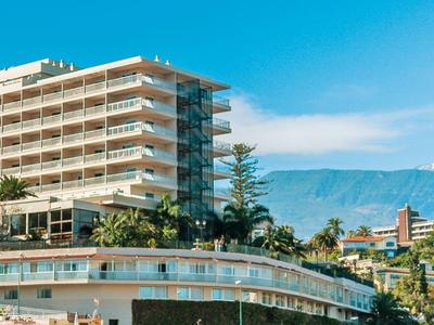Multi-story hotel with rows of balconies, surrounded by palm trees and mountains in the background.