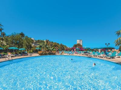 Großer runder Pool mit klarem Wasser, umgeben von Sonnenliegen, Palmen und blauem Himmel.