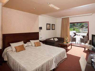 Helles Hotelzimmer mit großem Doppelbett, Sofa, TV und Terrasse mit Blick ins Grüne.
