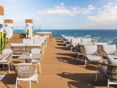 Dachterrasse mit weißen Stühlen, Holzboden, Pflanzen und Blick auf das blaue Meer unter klarem Himmel.