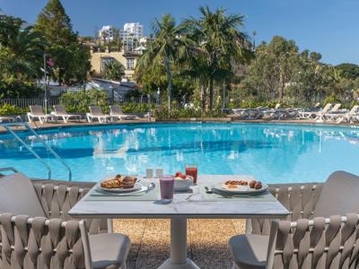 Tisch mit Getränken und Snacks am Pool eines Hotels mit Palmen und Liegestühlen.