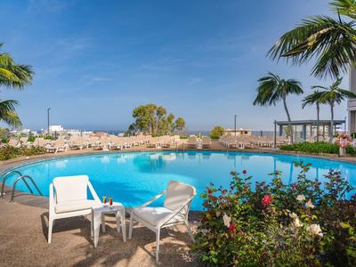 Runder Pool mit weißen Stühlen, Palmen und Meerblick an einem sonnigen Urlaubsort.